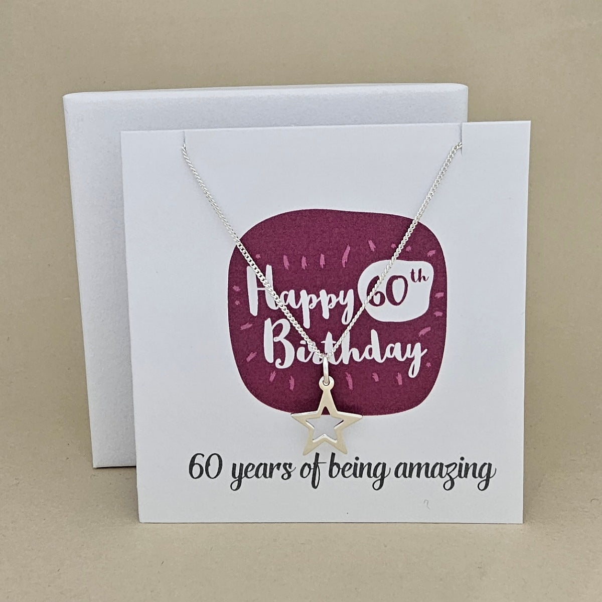 A silver open star necklace displayed on a white card with a burgundy rounded square and the text 'Happy 60th Birthday' and '60 years of being amazing,'.
