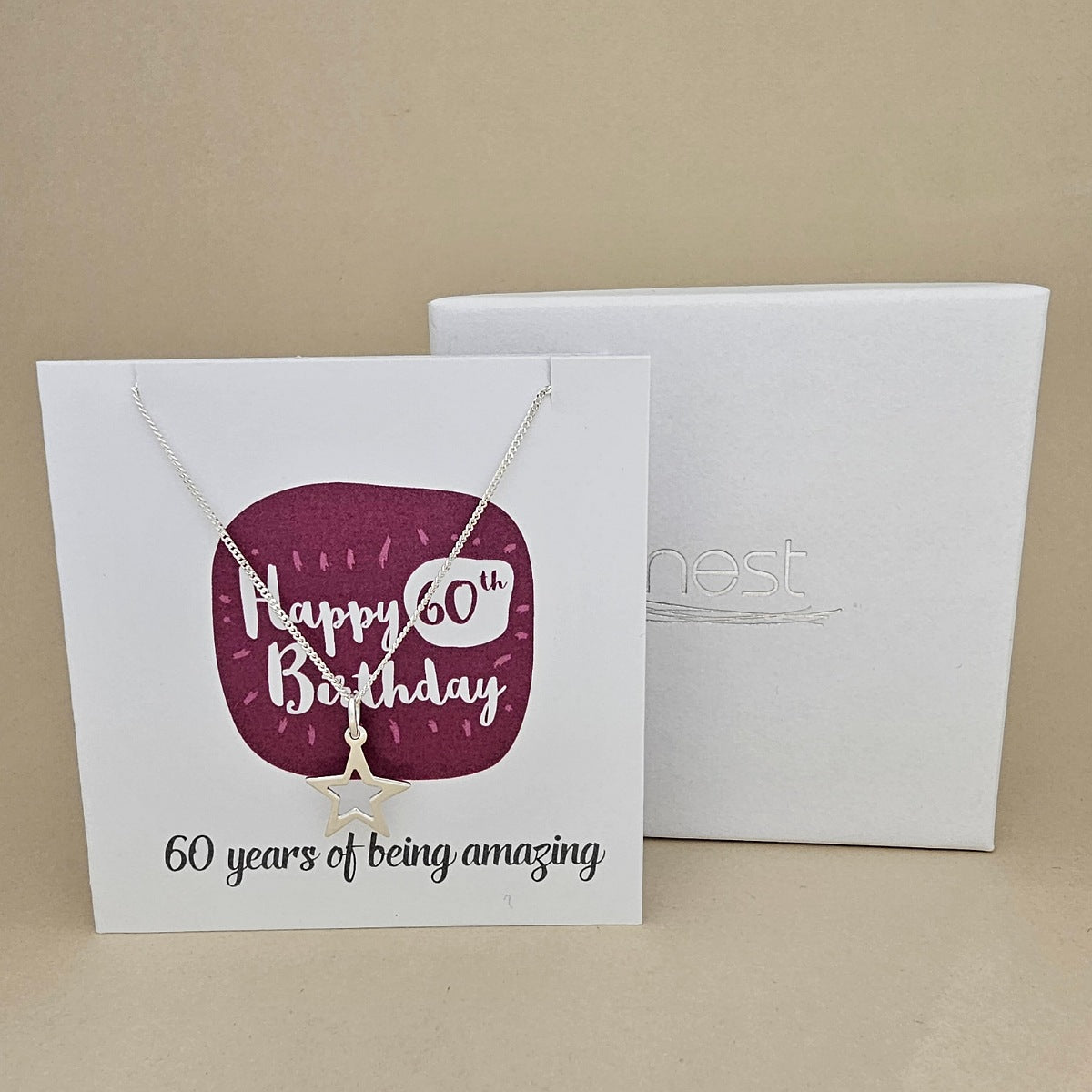 A silver open star necklace displayed on a white card with a burgundy rounded square and the text 'Happy 60th Birthday' and '60 years of being amazing,' shown in front of a white branded 'nest' gift box on a beige background.