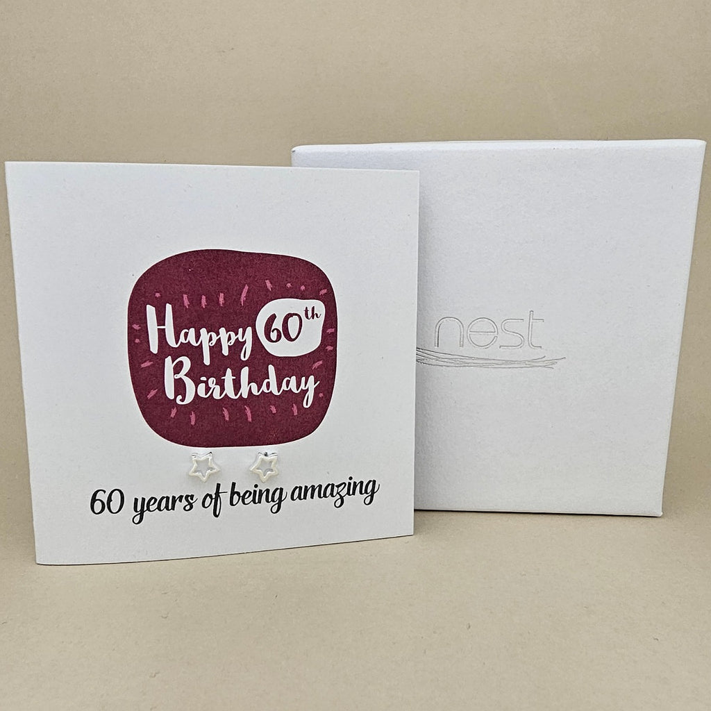 A pair of silver open star earrings displayed on a white card with a burgundy rounded square and the text 'Happy 60th Birthday' and '60 years of being amazing,' shown in front of a white branded 'nest' gift box on a beige background.