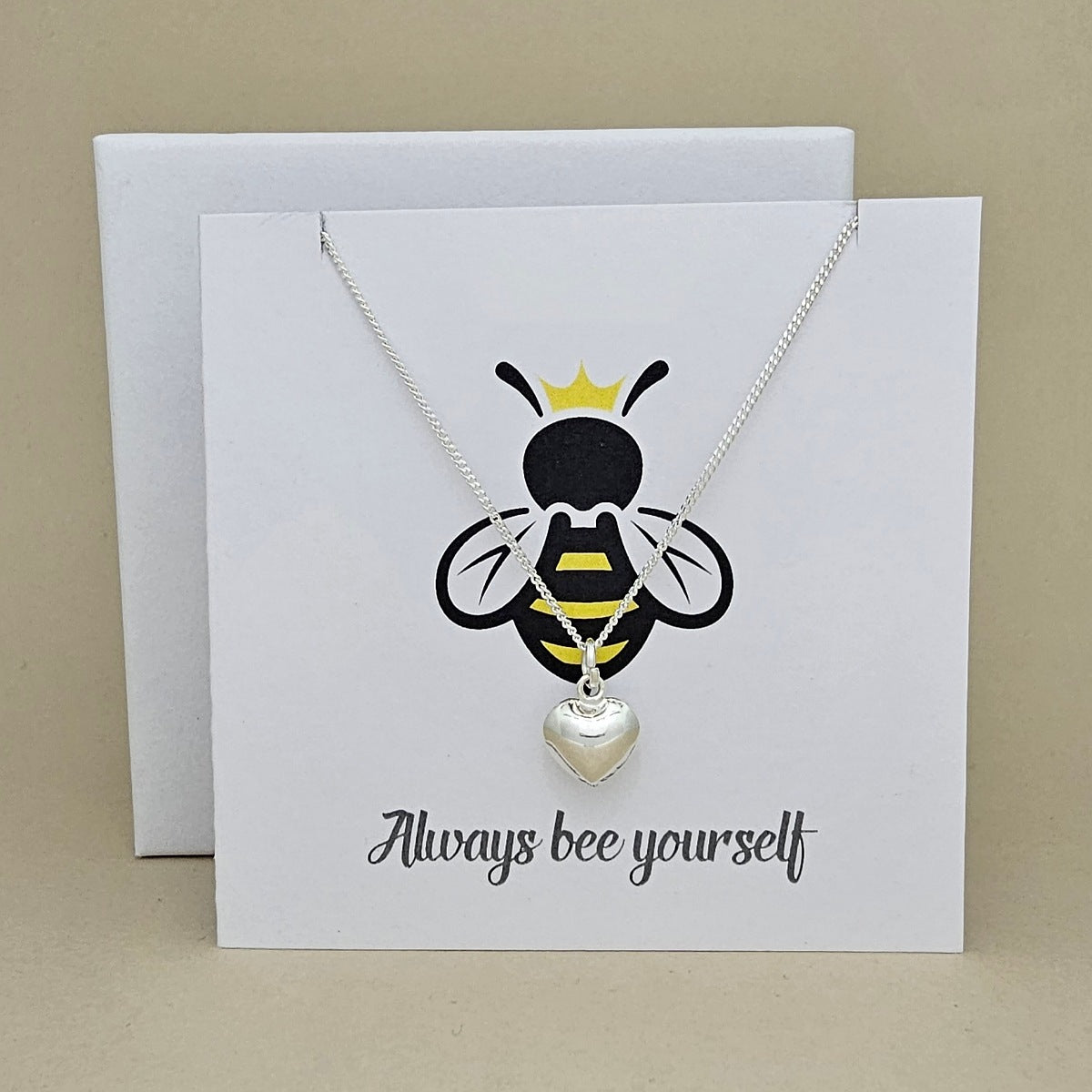 A silver necklace with a heart-shaped pendant is displayed on a white gift card featuring a bee illustration and the phrase 'Always bee yourself'.