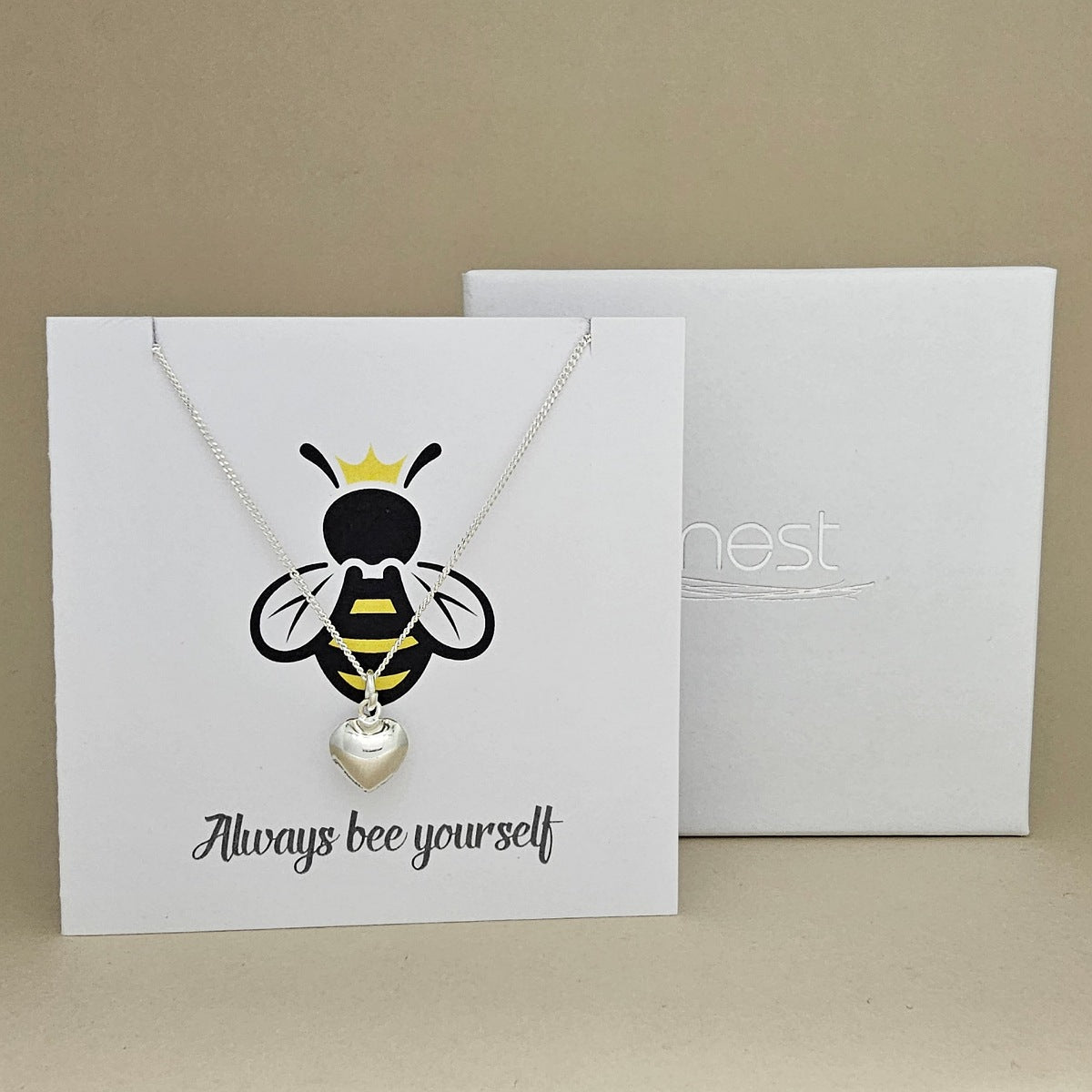 A silver necklace with a heart-shaped pendant is displayed on a white gift card featuring a bee illustration and the phrase 'Always bee yourself'.  Next to a white box with 'nest' branding