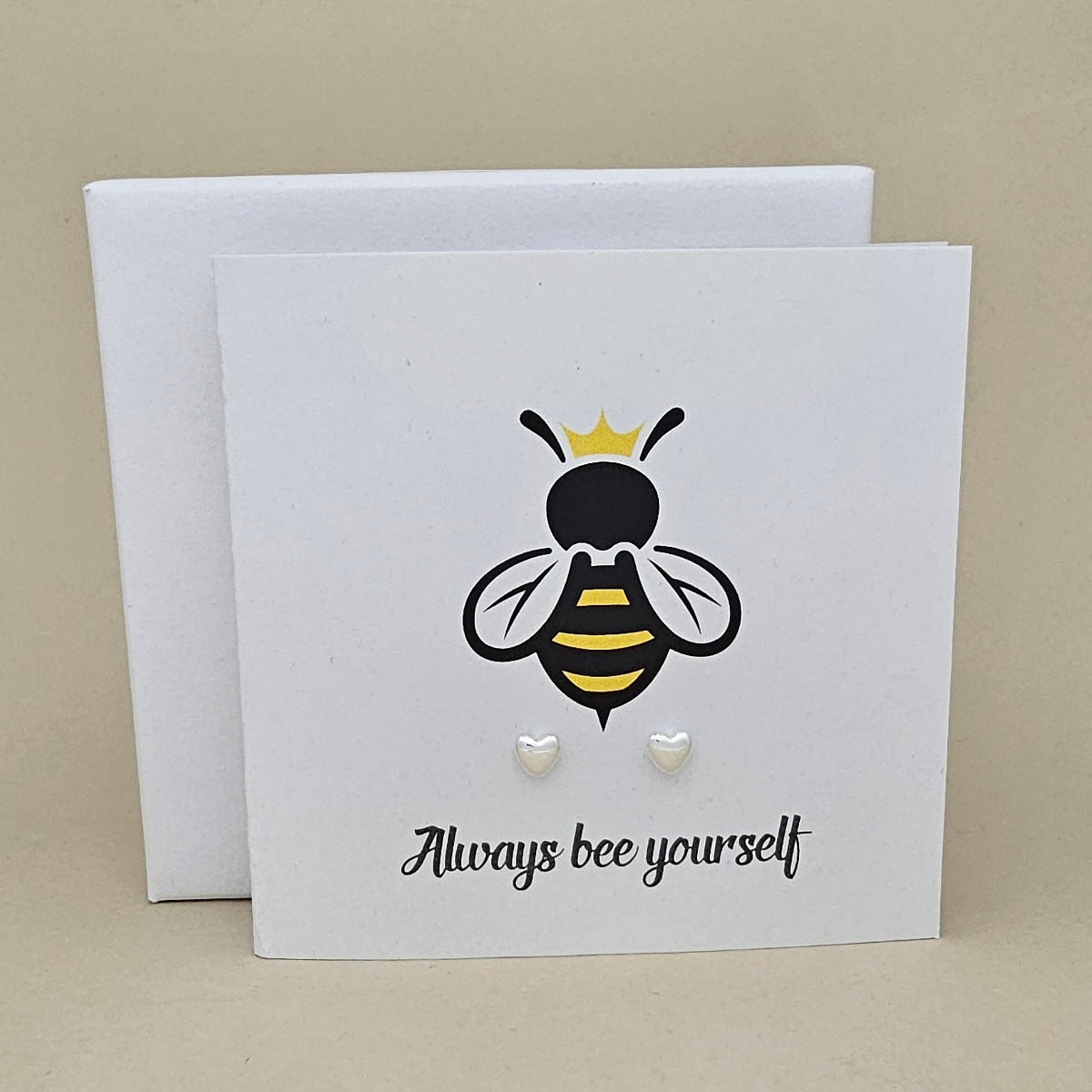A pair of silver heart shaped earrings are displayed on a white gift card featuring a bee illustration and the phrase 'Always bee yourself'.