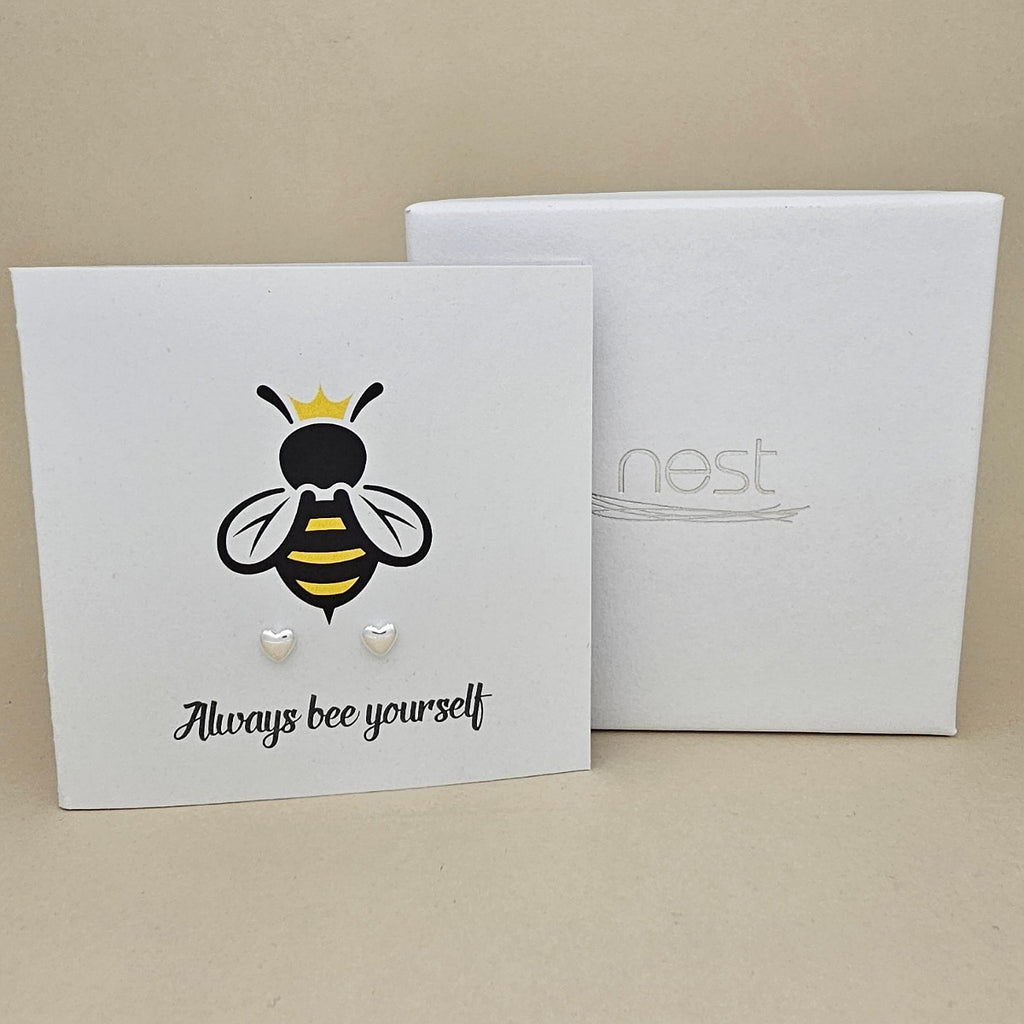 A pair of silver heart shaped earrings are displayed on a white gift card featuring a bee illustration and the phrase 'Always bee yourself'.  Next to a white box with 'nest' branding