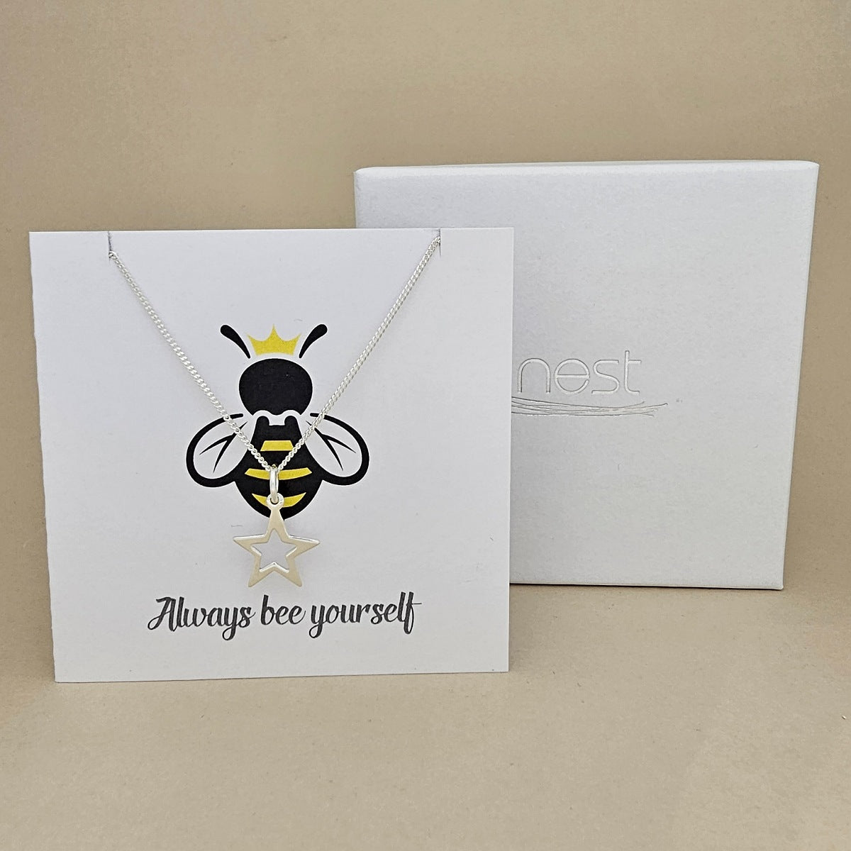 A silver necklace with an open star shaped pendant is displayed on a white gift card featuring a bee illustration and the phrase 'Always bee yourself'.  Next to a white box with 'nest' branding