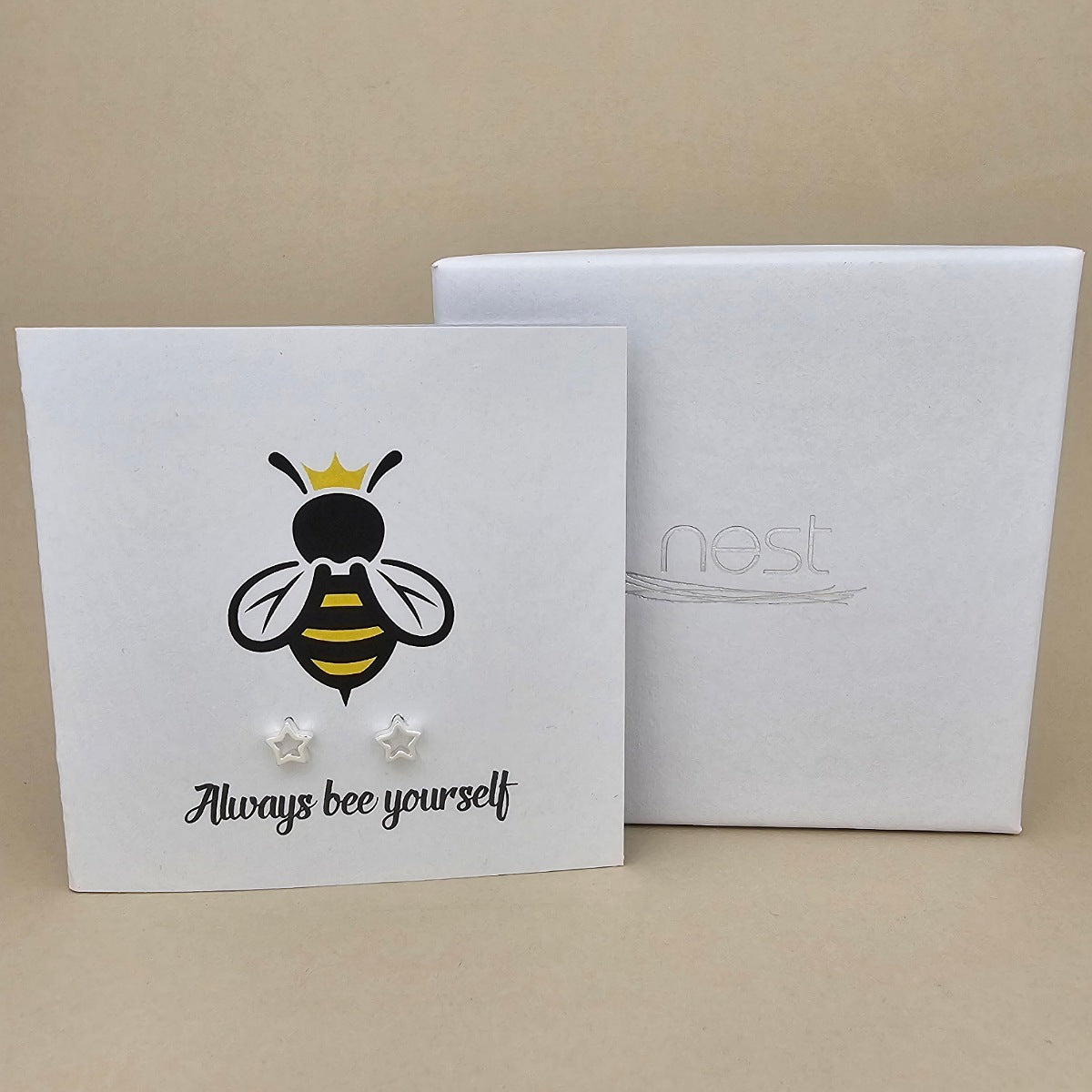 A pair of silver open star shaped earrings are displayed on a white gift card featuring a bee illustration and the phrase 'Always bee yourself'.  Next to a white box with 'nest' branding