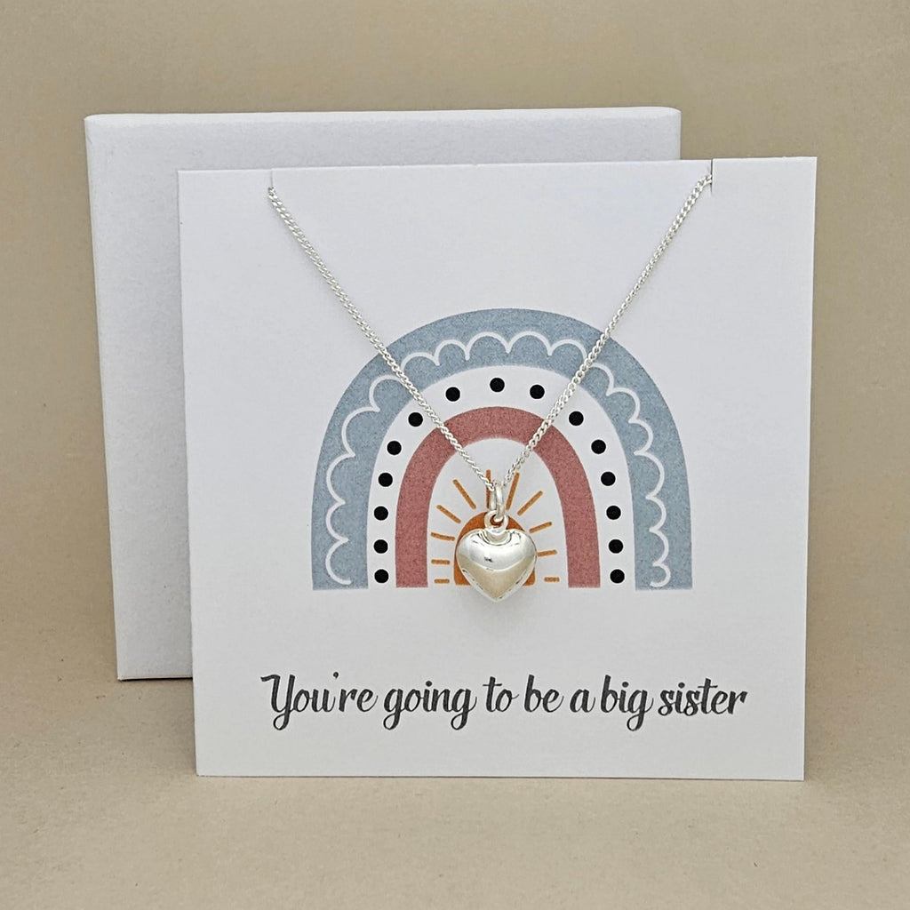 White square card with a silver necklace and heart pendant with rainbow design and text 'You're going to be a big sister' on a beige background.
