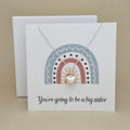 White square card with a silver necklace and heart pendant with rainbow design and text 'You're going to be a big sister' on a beige background.

