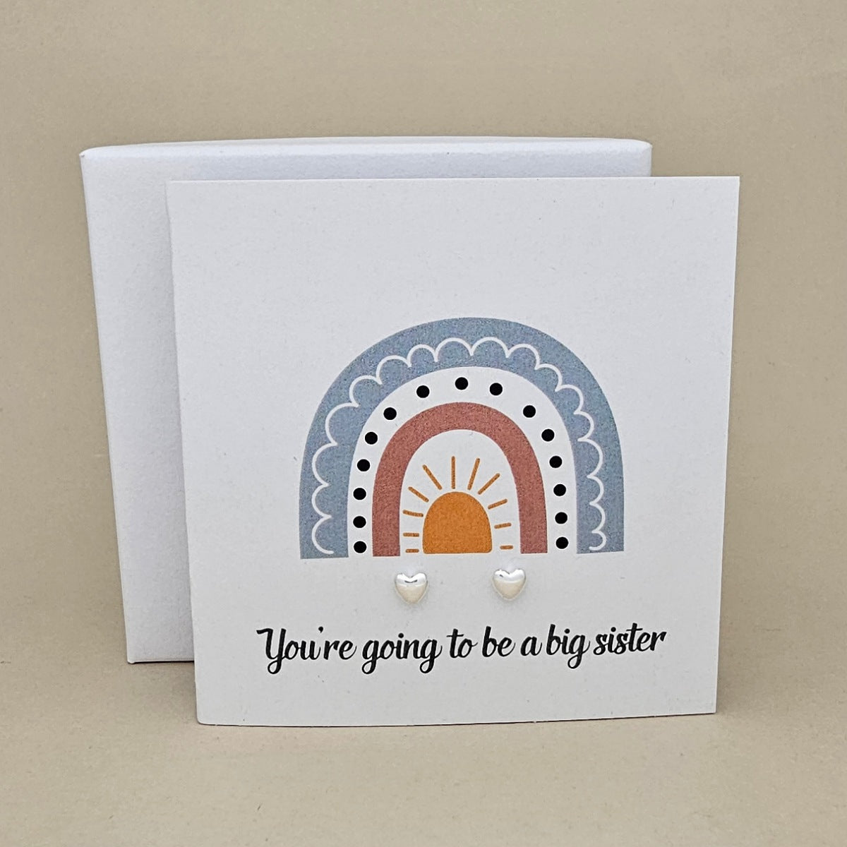 White square card with a pair of silver heart earrings with rainbow design and text 'You're going to be a big sister' on a beige background.   
