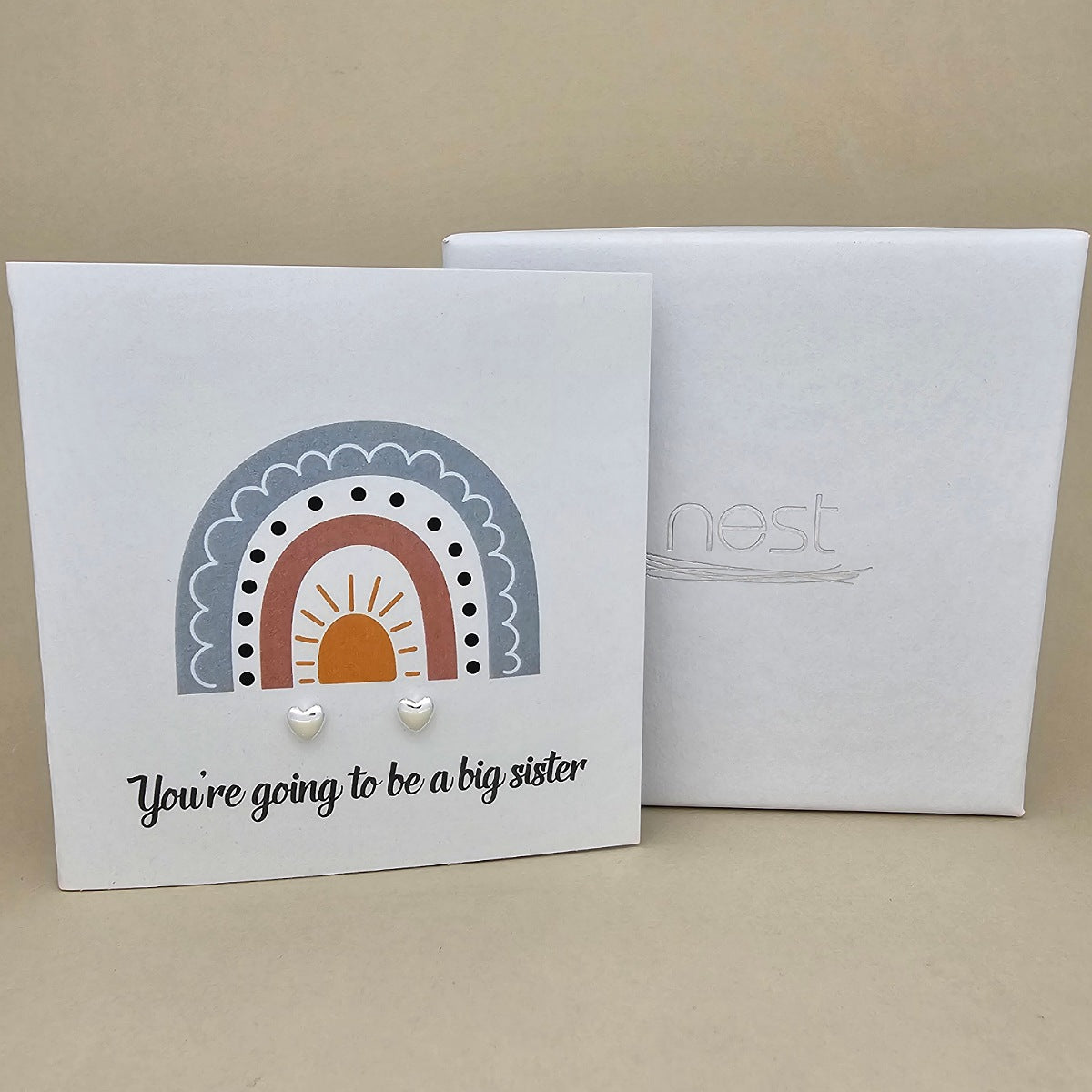 White square card with a pair of silver heart earrings with rainbow design and text 'You're going to be a big sister' on a beige background.   Next to a white box with 'nest' branding
