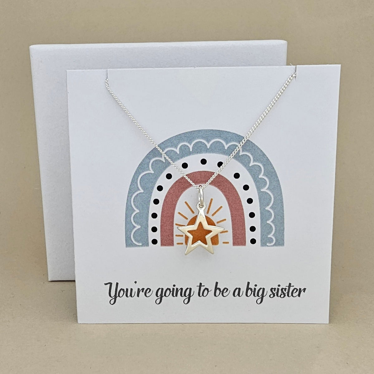 White square card with a silver necklace and open star pendant with rainbow design and text 'You're going to be a big sister' on a beige background.
