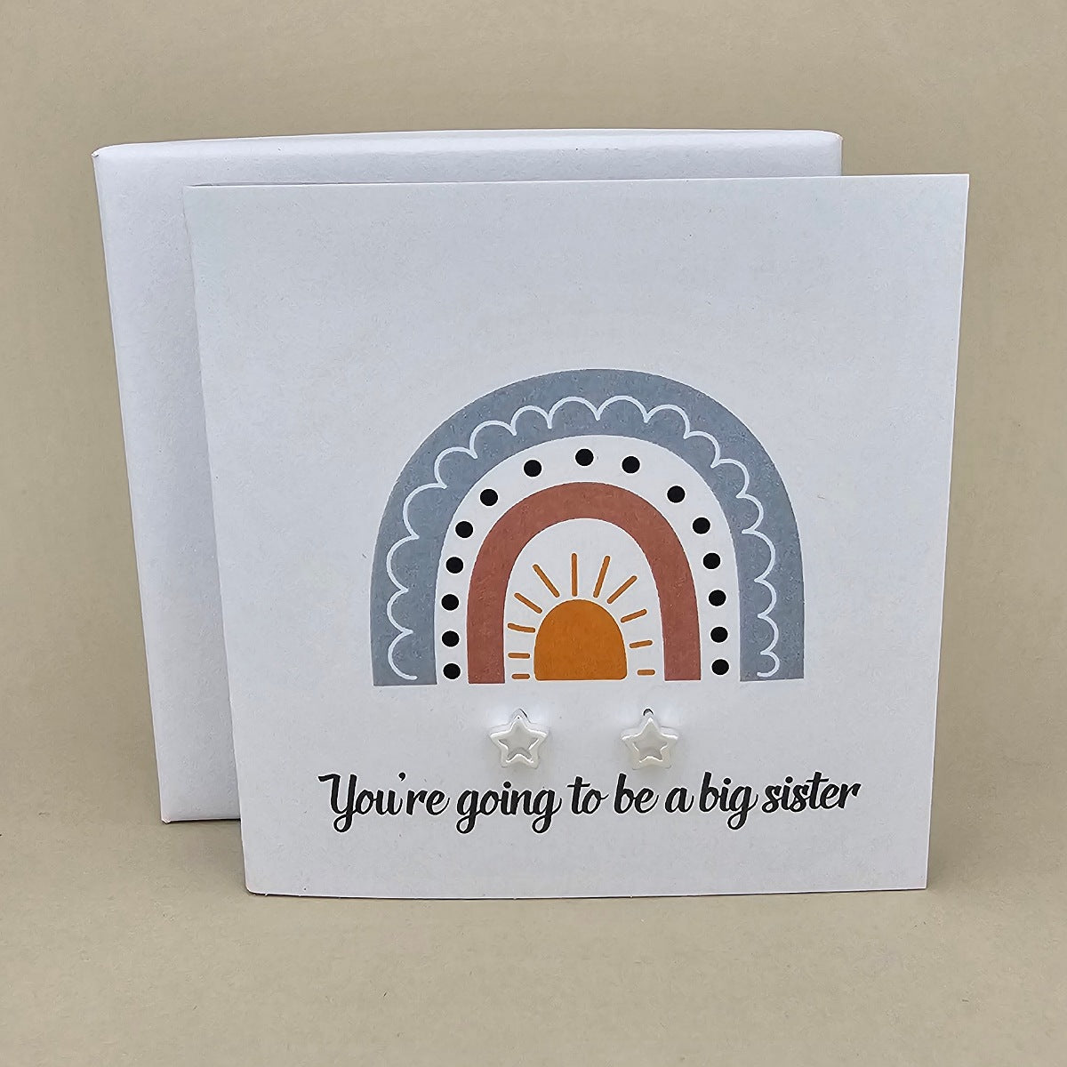 White square card with a pair of silver open star earrings with rainbow design and text 'You're going to be a big sister' on a beige background