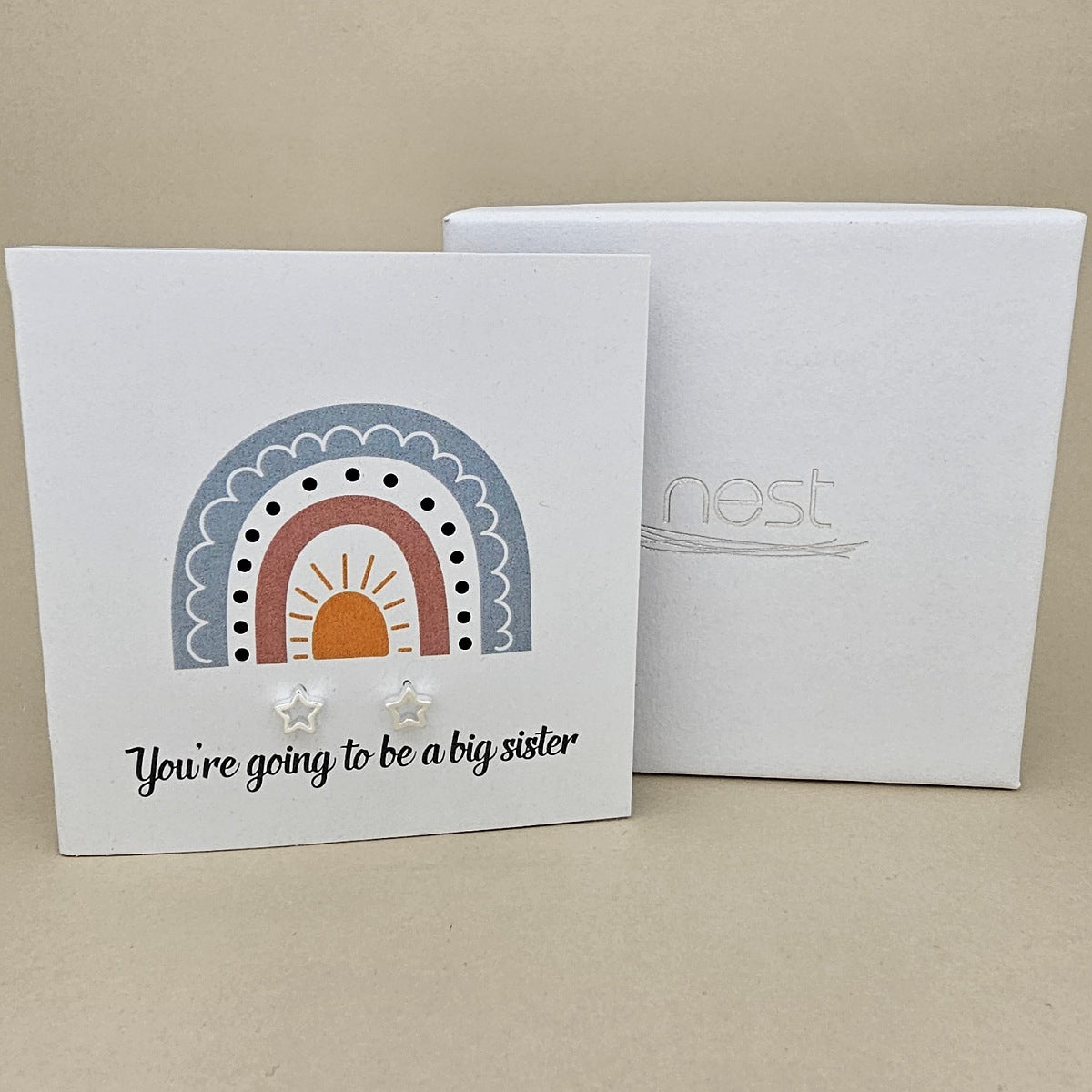 White square card with a pair of silver open star earrings with rainbow design and text 'You're going to be a big sister' on a beige background.   Next to a white box with 'nest' branding
