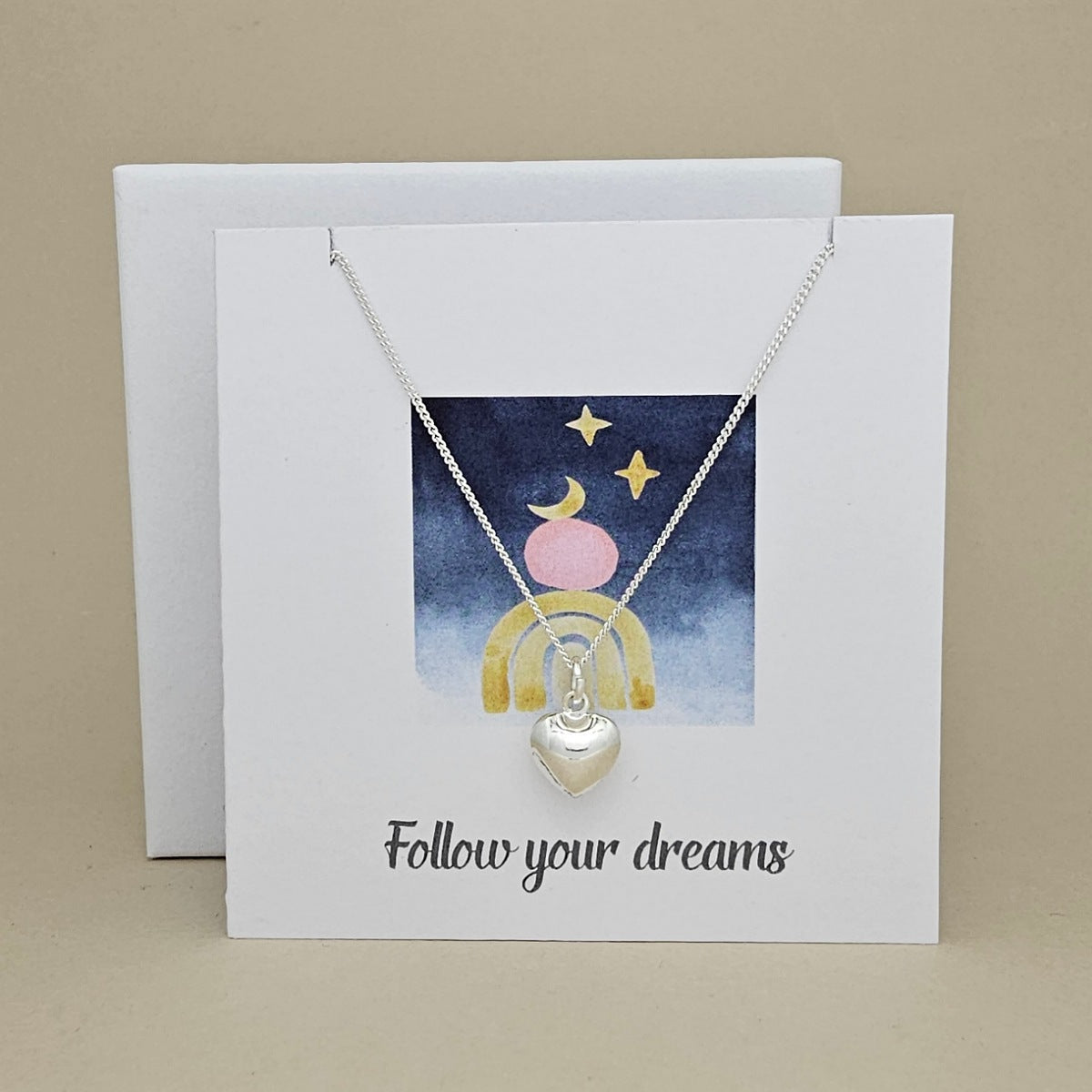 Silver necklace with heart pendant on a card with 'Follow your dreams' text and celestial design.