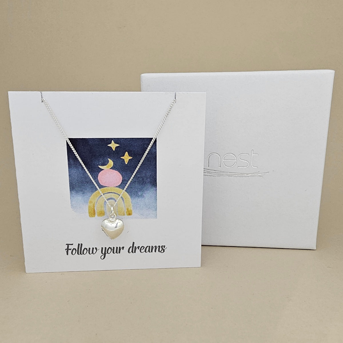 Silver necklace with heart pendant on a card with 'Follow your dreams' text and celestial design.  Next to a white box with 'nest' branding.