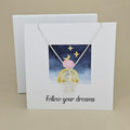 White card with a celestial design and a silver necklace with an open star pendant with the text 'Follow your dreams' on a beige background, next to a white box.