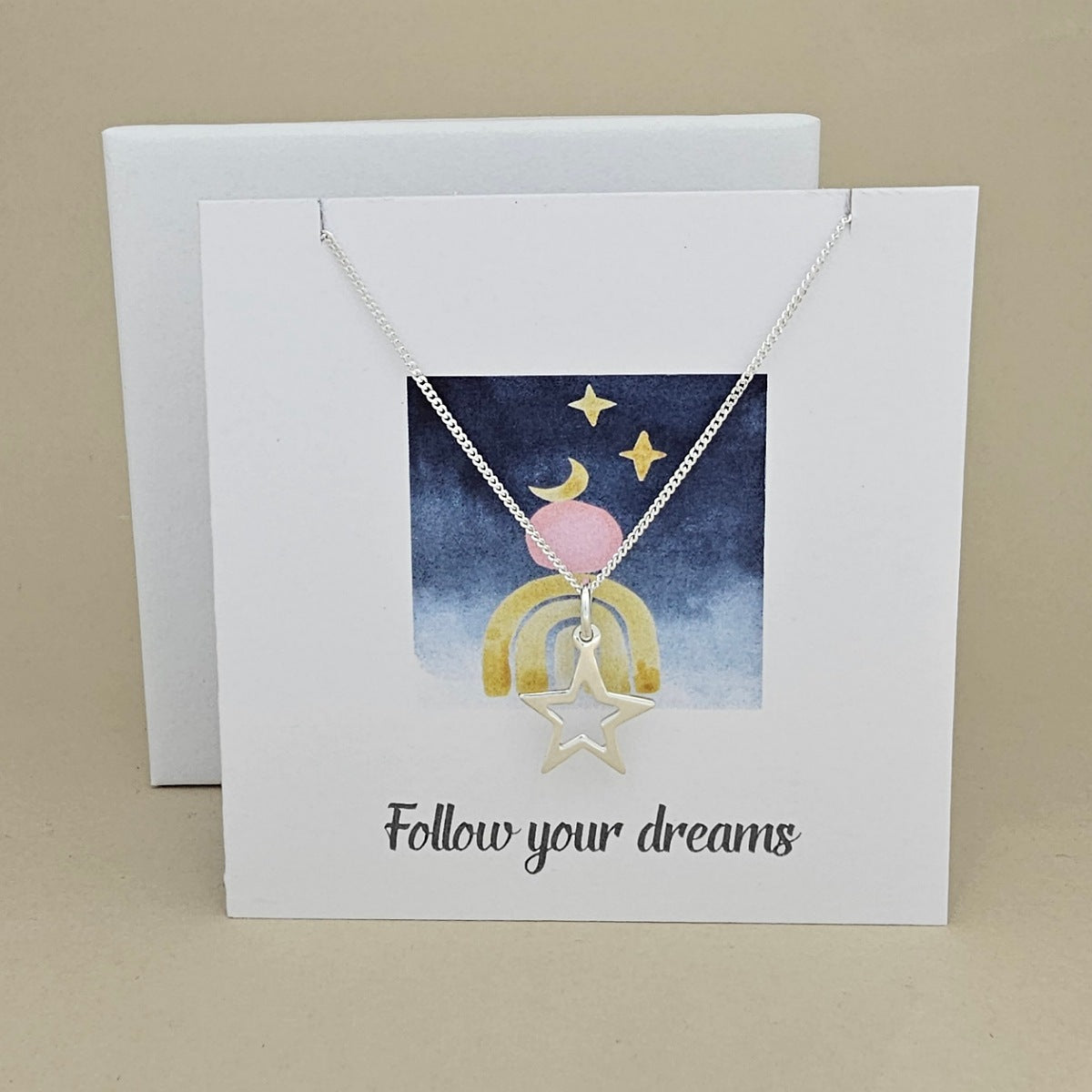 White card with a celestial design and a silver necklace with an open star pendant with the text 'Follow your dreams' on a beige background, next to a white box.