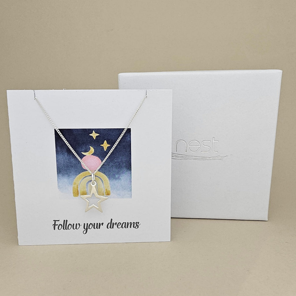 White card with a celestial design and a silver necklace with an open star pendant with the text 'Follow your dreams' on a beige background, next to a white box with 'nest' branding.