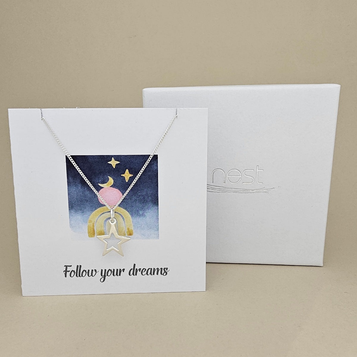 White card with a celestial design and a silver necklace with an open star pendant with the text 'Follow your dreams' on a beige background, next to a white box with 'nest' branding.