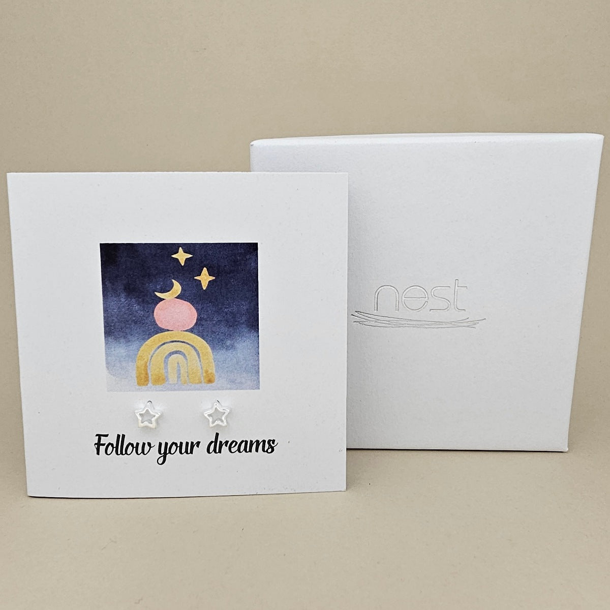 White card with a celestial design and a pair of silver open star earrings and 'Follow your dreams' text on a beige background, next to a white box with 'nest' branding.