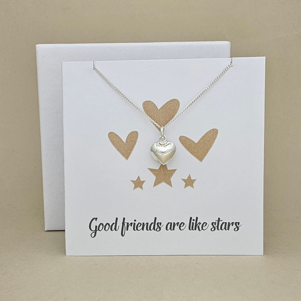 A silver heart pendant necklace on a delicate chain, displayed on a white card that reads 'Good friends are like stars' with gold heart and star graphics, set against a beige background.