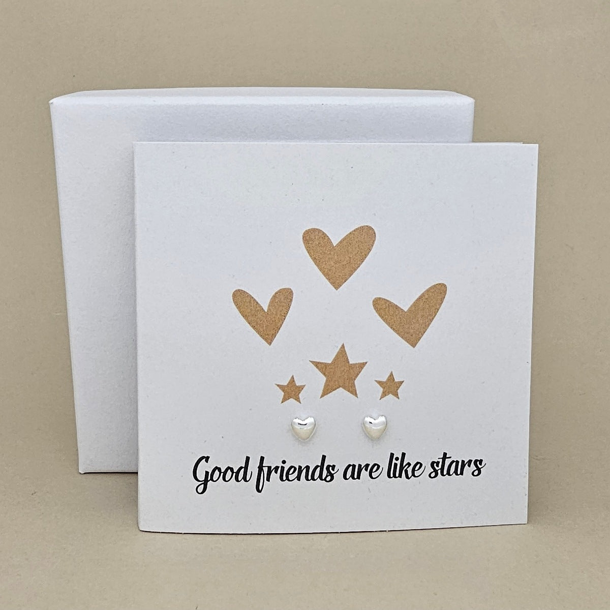 A white square card with the phrase 'Good friends are like stars' in black script, decorated with gold hearts and stars.  Two silver small heart stud earrings. A white gift box is placed behind the card on a beige background.