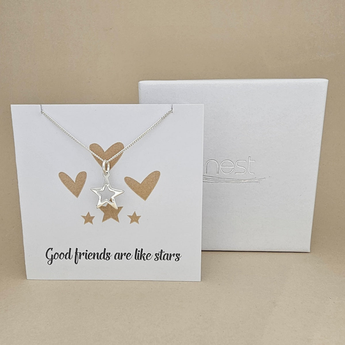 White card with gold hearts and stars design and 'Good friends are like stars' text, hanging silver necklace and open star pendant next to a white box with 'nest' branding.
