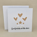A square white greeting card with the phrase 'Good friends are like stars' in black script, decorated with gold hearts, stars, and two silver small open star earrings, shown in front of a matching white box on a beige background.