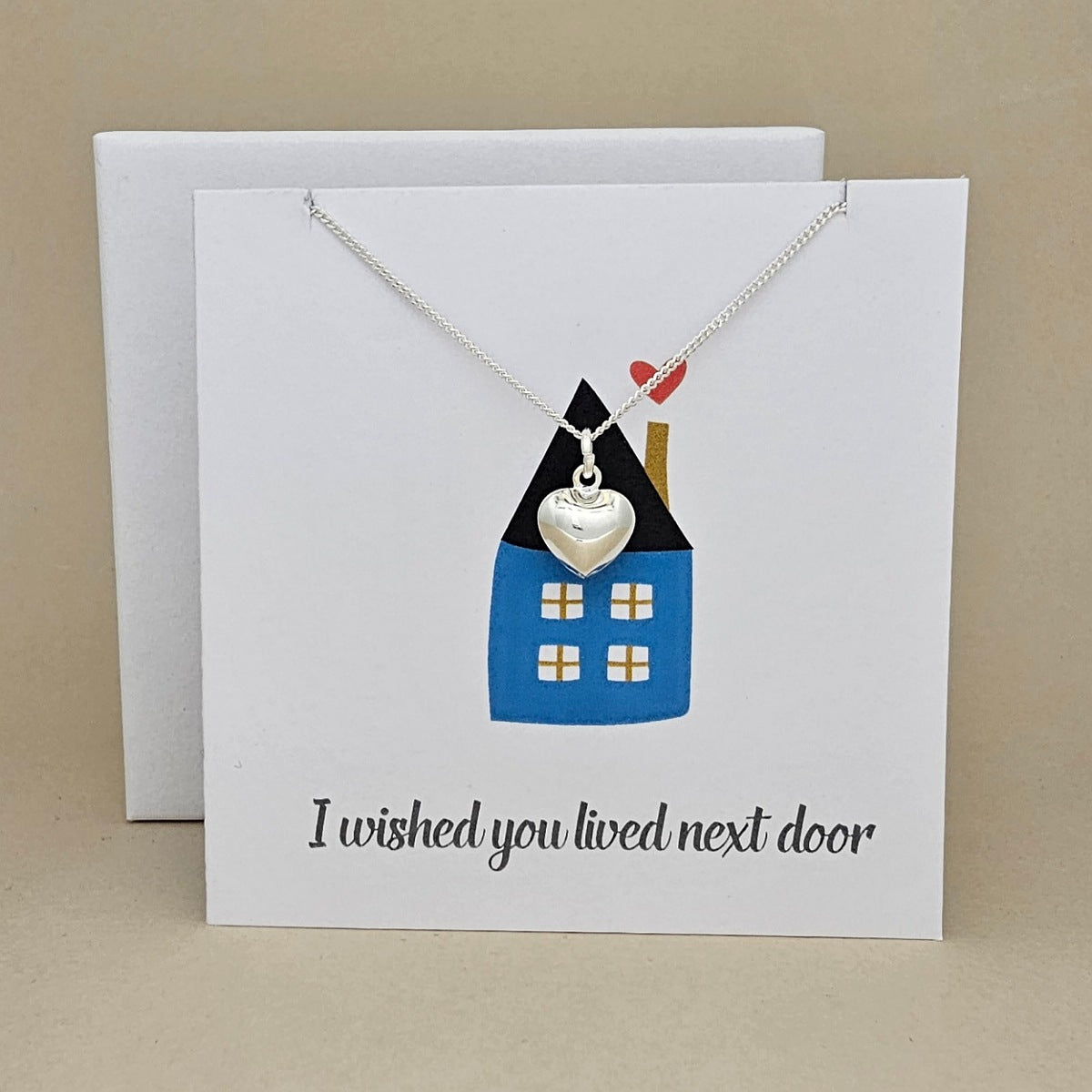 White square card with a silver necklace and heart pendant on a card featuring a house illustration and text 'I wished you lived next door' against a beige background.