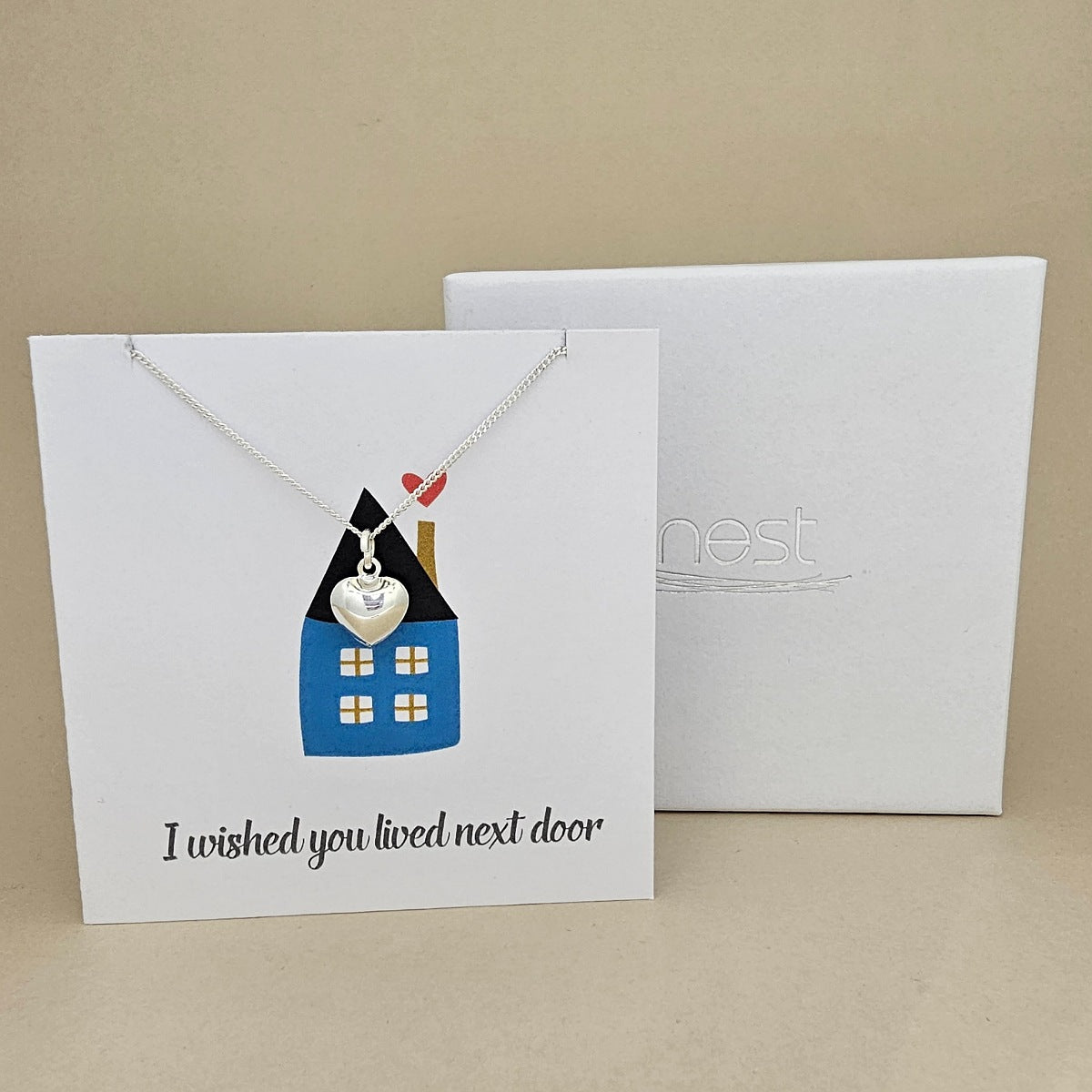 Silver necklace with a  heart pendant on a card with 'I wished you lived next door' text and house image, displayed against a beige background.