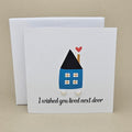 A white greeting card with a small illustrated house in blue, black, and red, plus the text 'I wished you lived next door' and two small silver heart earrings, shown with a white box behind it on a beige background.

