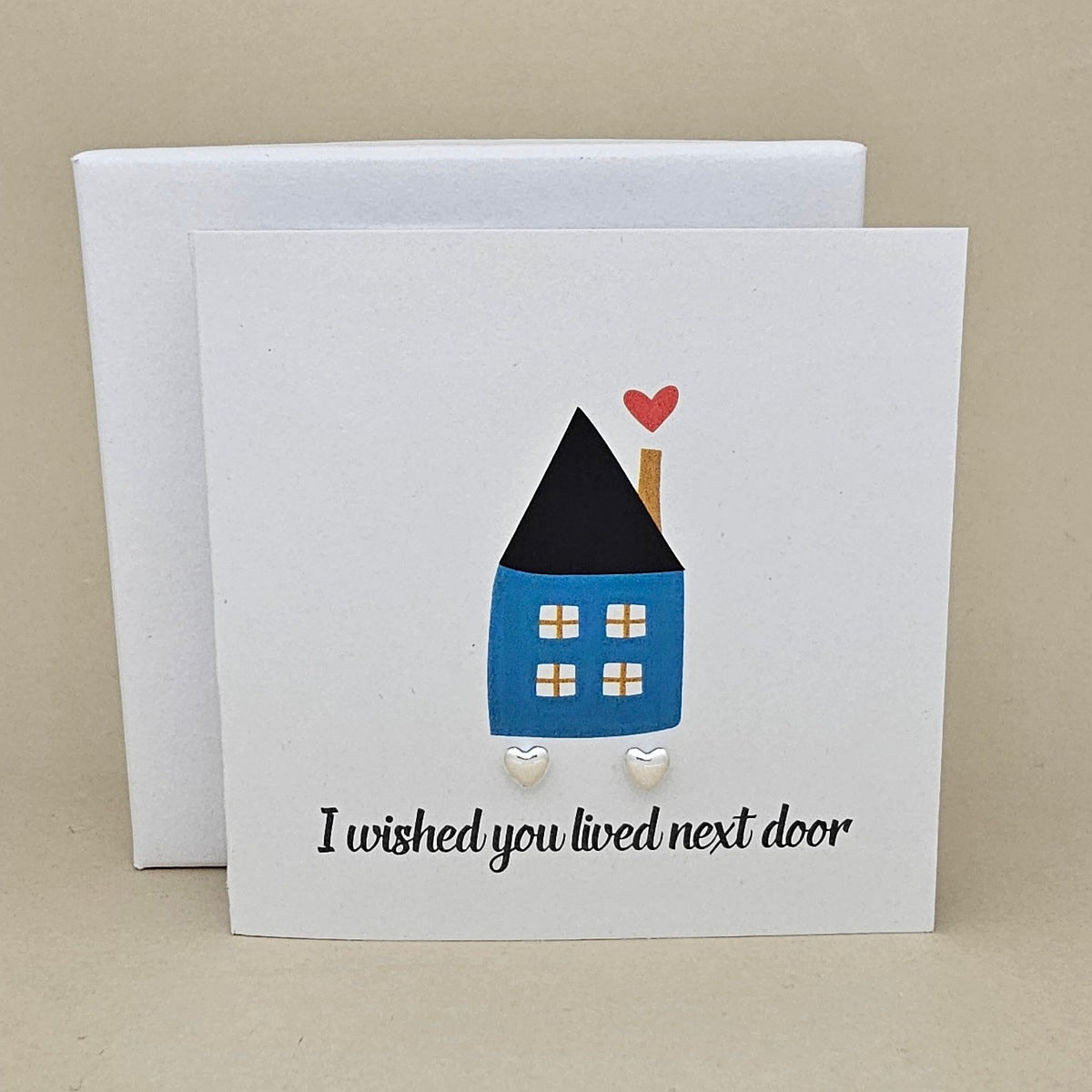 A white greeting card with a small illustrated house in blue, black, and red, plus the text 'I wished you lived next door' and two small silver heart earrings, shown with a white box behind it on a beige background.

