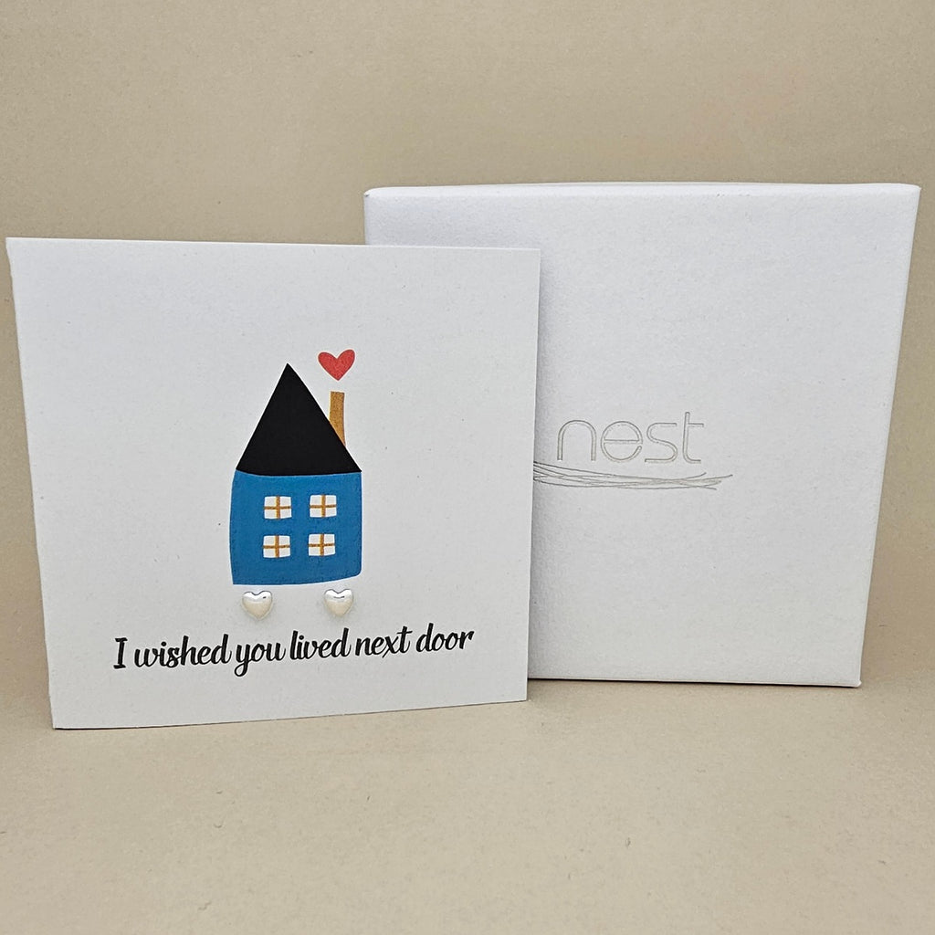 Square card with a house illustration and two silver heart earrings with the text 'I wished you lived next door' next to a white box on a beige background.