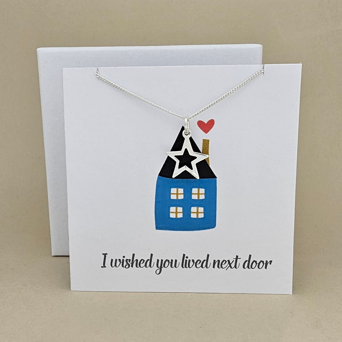 A silver necklace with a open star pendant, displayed on a white greeting card that reads 'I wished you lived next door' and an image of a house against a neutral beige background.