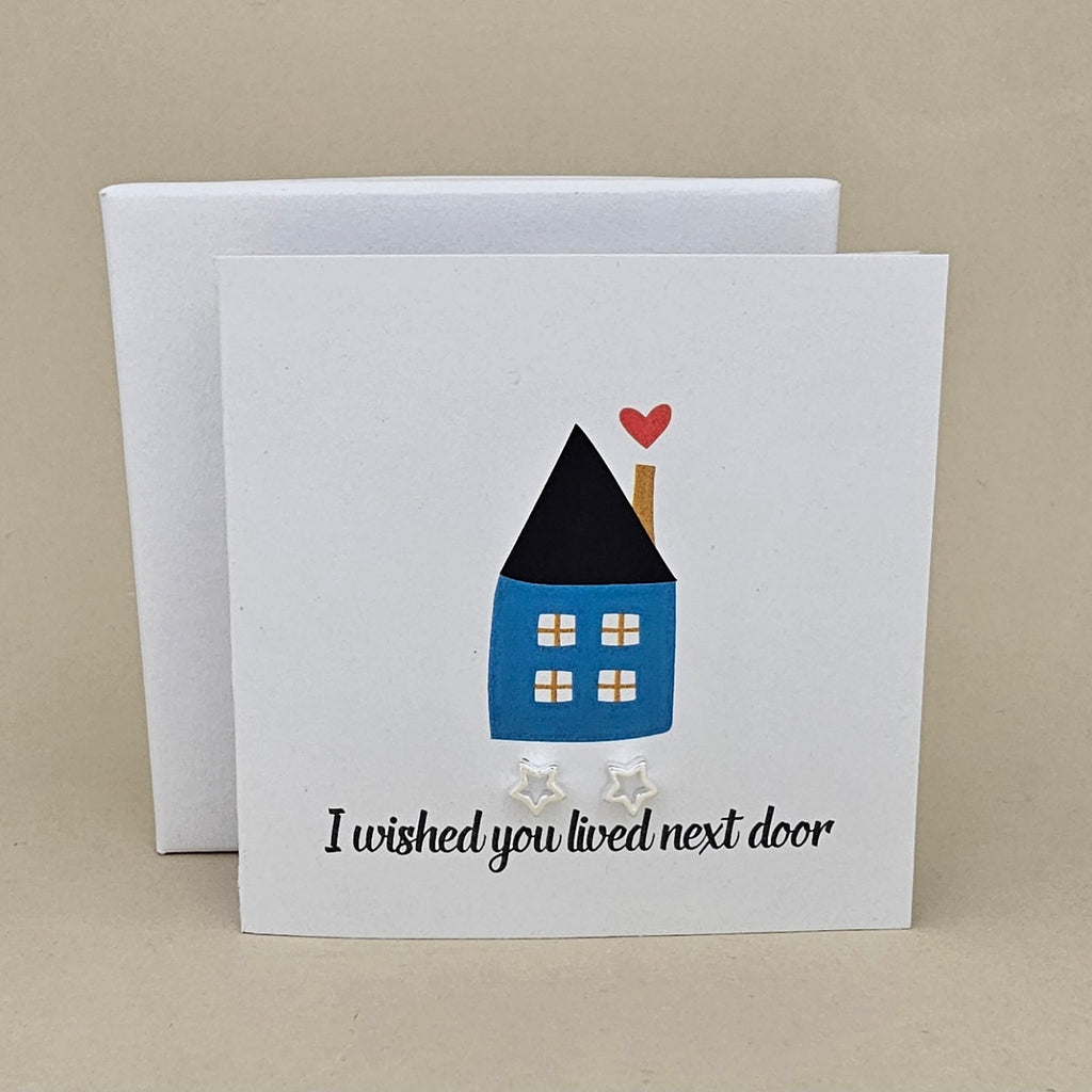 A white greeting card with a small illustrated house in blue, black, and red, plus the text 'I wished you lived next door' and two small silver open star earrings, shown with a white box behind it on a beige background.