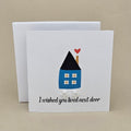 A white greeting card with a small illustrated house in blue, black, and red, plus the text 'I wished you lived next door' and two small silver open star earrings, shown with a white box behind it on a beige background.