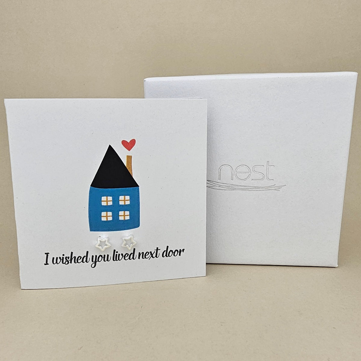 Square card with a house illustration and two silver open star earrings with the text 'I wished you lived next door' next to a white box on a beige background.