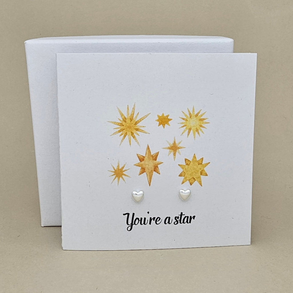 A white square card with gold star illustrations, the phrase 'You're a star' in black script, and two silver small heart-shaped stud earrings displayed on it, with a white box behind it on a beige background.