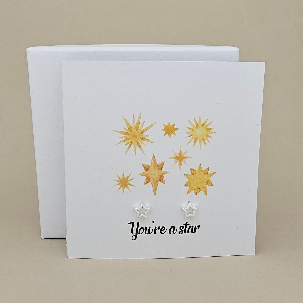 A square white greeting card with gold and white star illustrations and a pair of silver open star earrings, with printed wording 'You're a star' in black script, shown in front of a matching white box on a warm beige background.