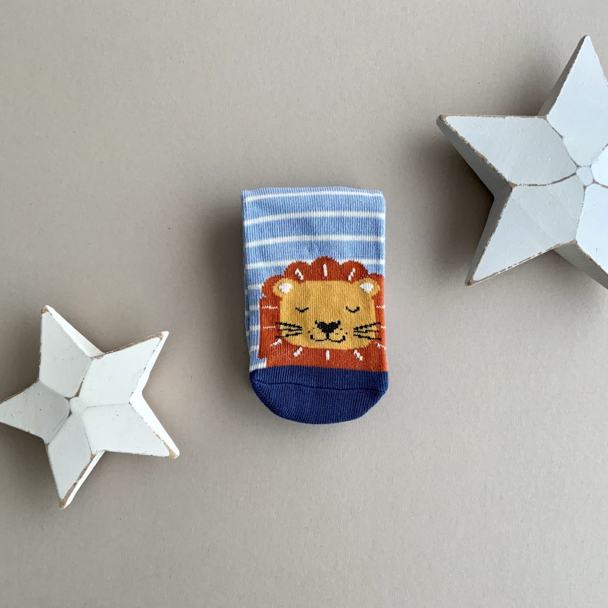 A single folded blue and white striped sock with an orange and yellow lions head.   Featuring a navy cuff and toe, displayed on a neutral beige background with white star-shaped props.

