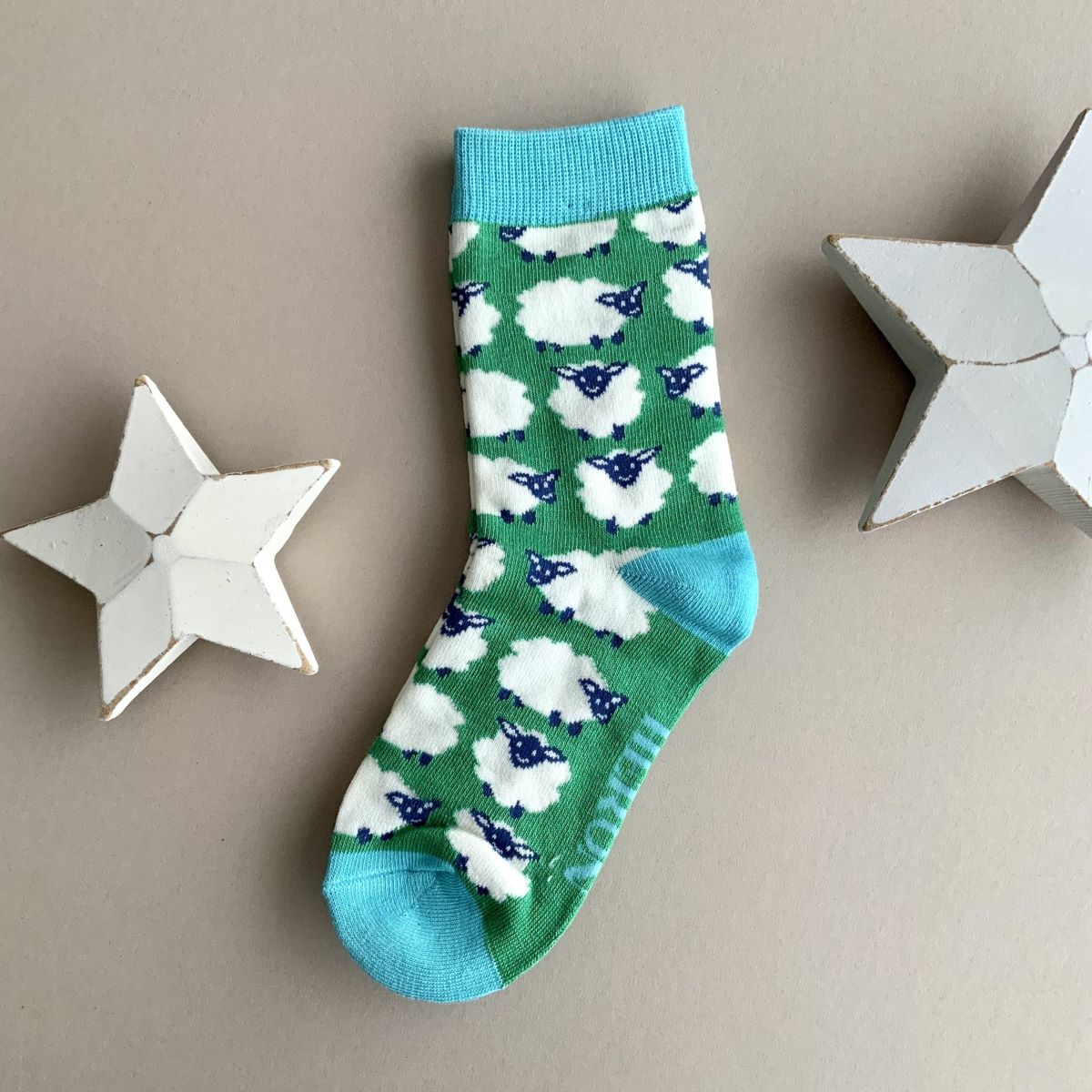 A single green sock with a repeating pattern of white sheep with navy faces.   Featuring a light blue cuff, heal and toe, displayed on a neutral beige background with white star-shaped props.
