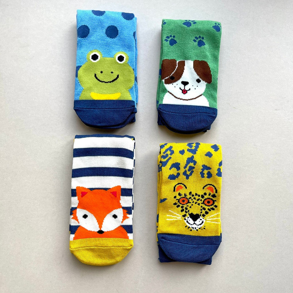 Four brightly coloured children's socks laid folded flat on a light beige background, each with a different animal face design: a dog on a green sock with blue paw prints, a green frog on a blue sock with navy spots, a yellow leopard with navy print, and a orange fox on a navy-and-white stripe sock.