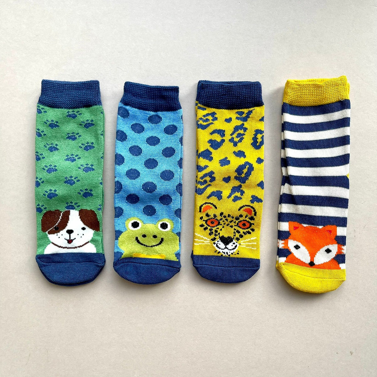 Four brightly coloured children's socks laid flat on a light beige background, each with a different animal face design: a dog on a green sock with blue paw prints, a green frog on a blue sock with navy spots, a yellow leopard with navy print, and a orange fox on a navy-and-white stripe sock.