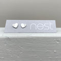 A pair of small silver heart stud earrings displayed on a gray branded card that reads 'nest', placed on a textured light gray surface against a soft gray background.