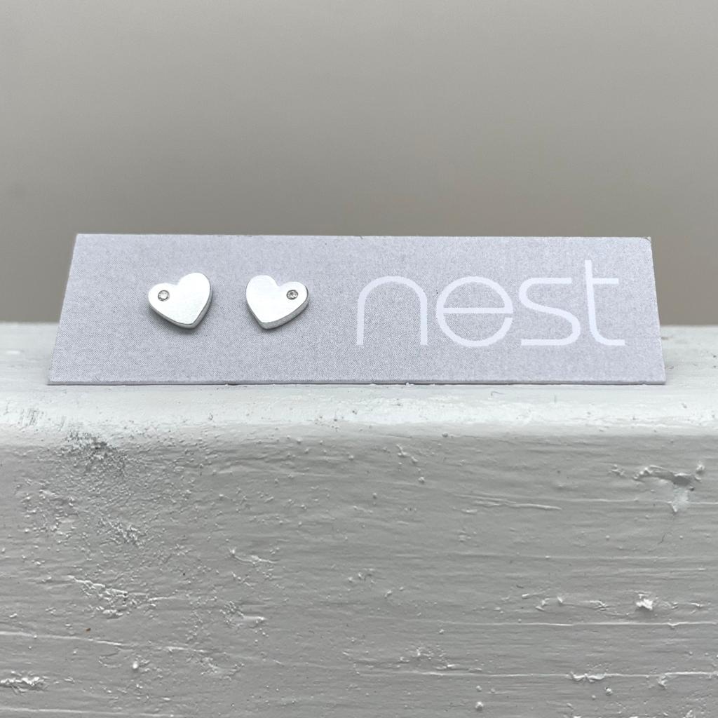 A pair of small silver heart stud earrings displayed on a gray branded card that reads 'nest', placed on a textured light gray surface against a soft gray background.