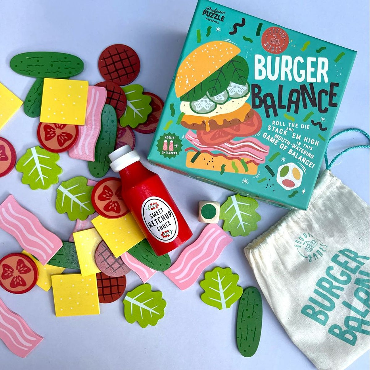 Burger Balance toy puzzle set with a turquoise game box, wooden cutout food pieces, a small red bottle, and a white drawstring bag on a pale blue background.