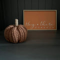 A chunky knit wool decorative pumpkin in a dusty burnt-orange colour with a natural jute stem, placed next to a framed sign that reads 'Hey there PUMPKIN' on a dark grey background.
