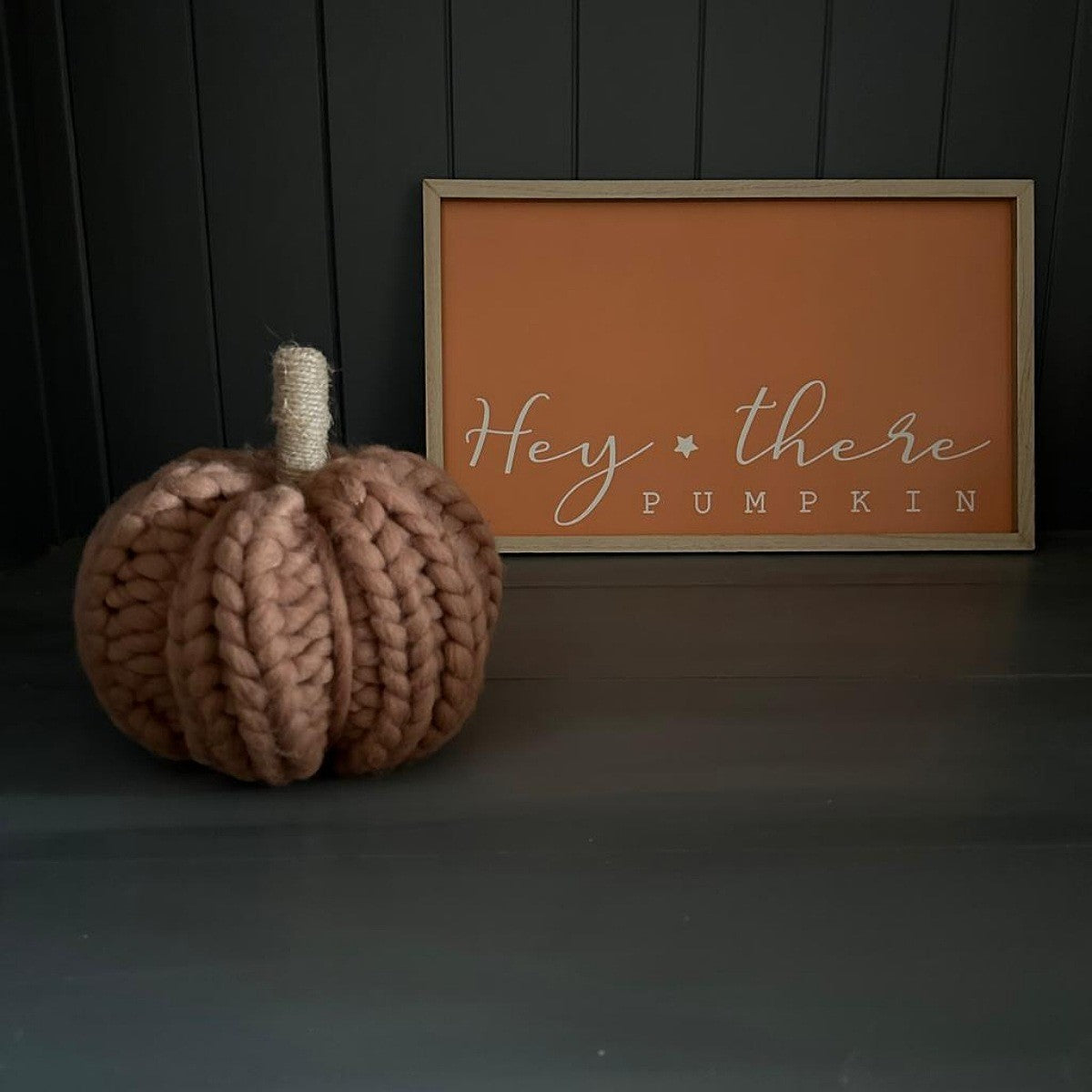 A chunky knit wool decorative pumpkin in a dusty burnt-orange colour with a natural jute stem, placed next to a framed sign that reads 'Hey there PUMPKIN' on a dark grey background.