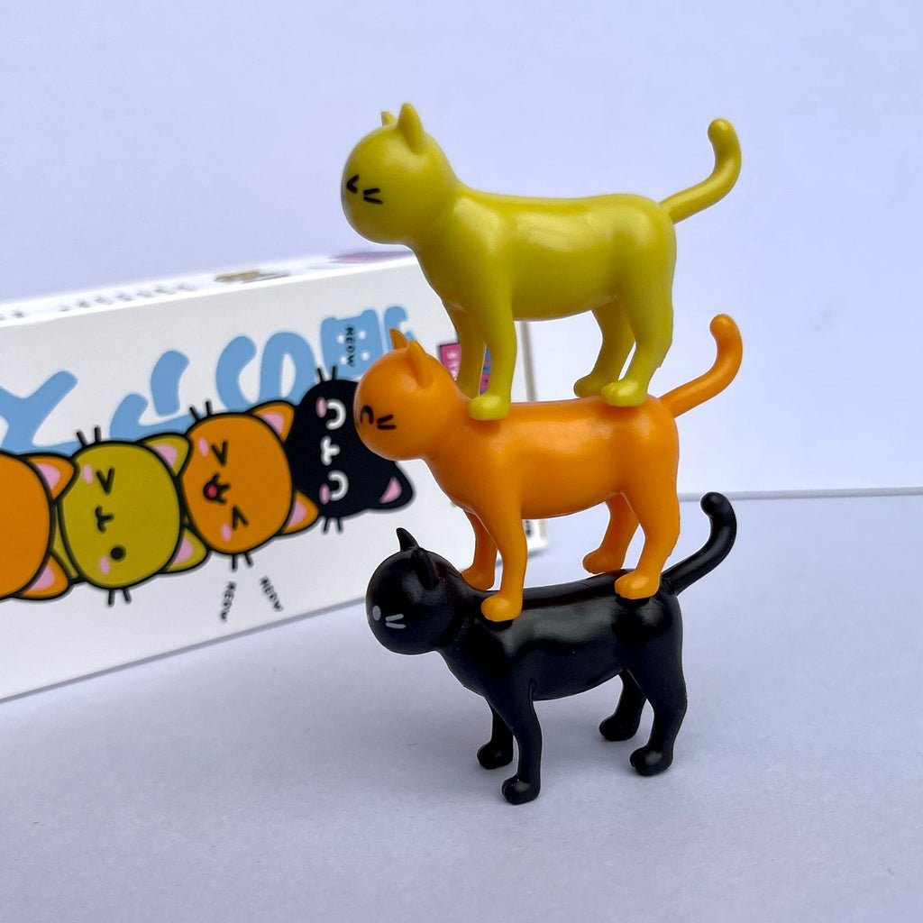 Three cat figurines stacked on top of each other with a colorful box in the background.