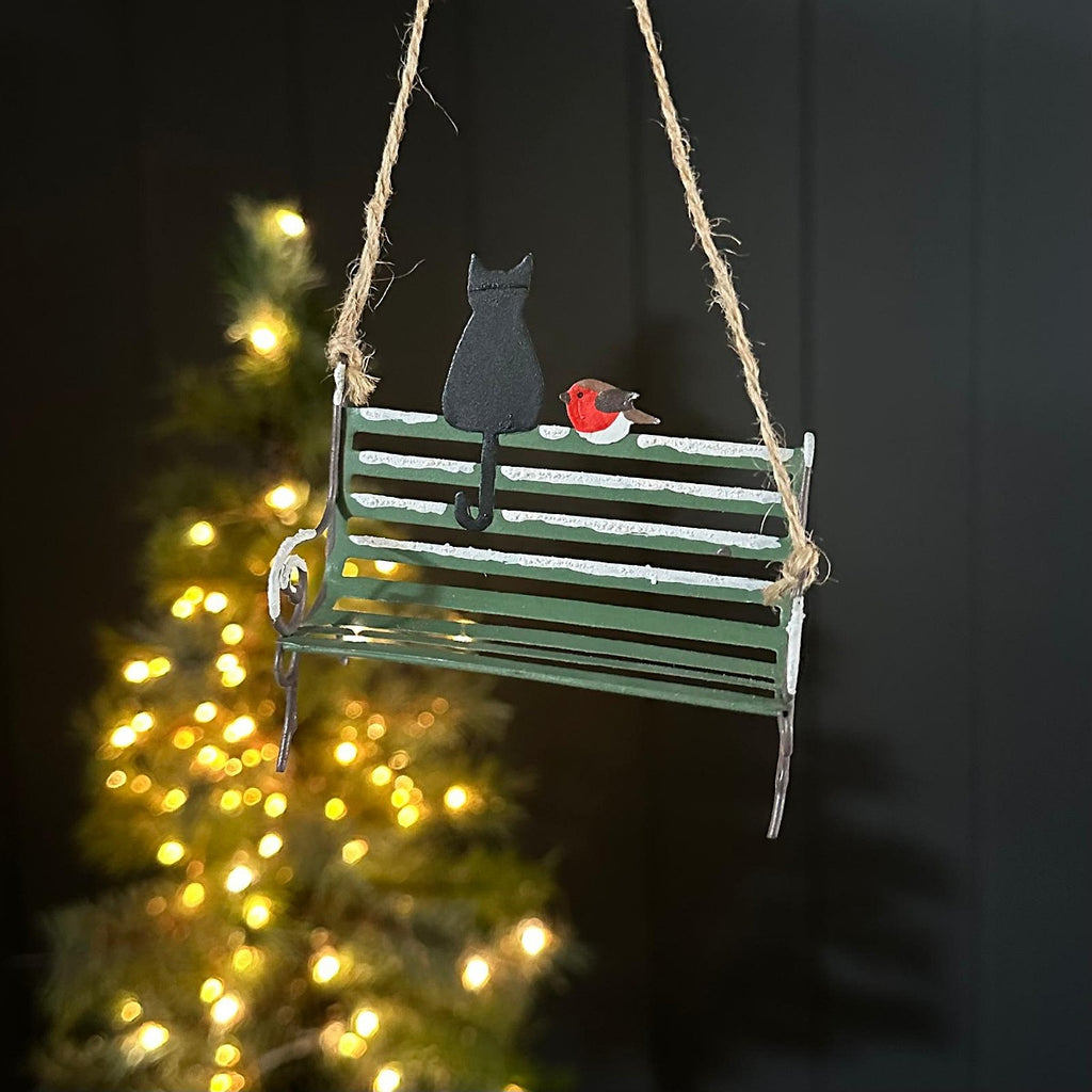 A hanging Christmas ornament shaped like a green wooden bench with a black cat silhouette and a red robin figure, suspended by twine against a dark background with blurred warm lights.
