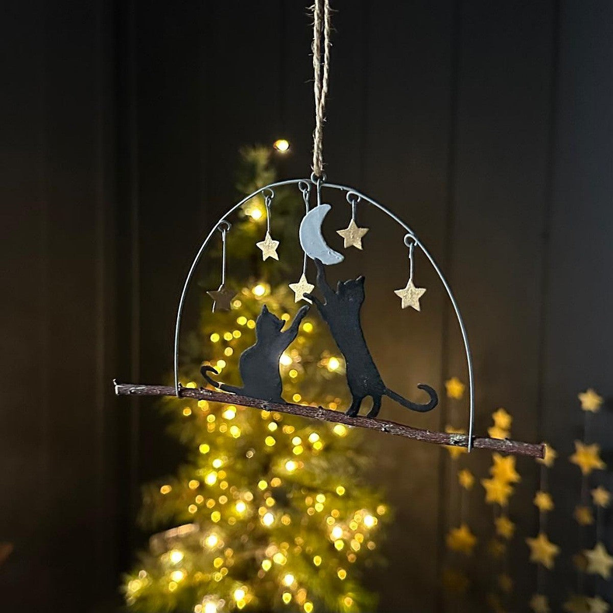 A hanging metal ornament shaped like two black cats reaching toward a crescent moon and stars, suspended from a twig branch with a twine string, shown in front of a softly blurred Christmas tree with warm lights.